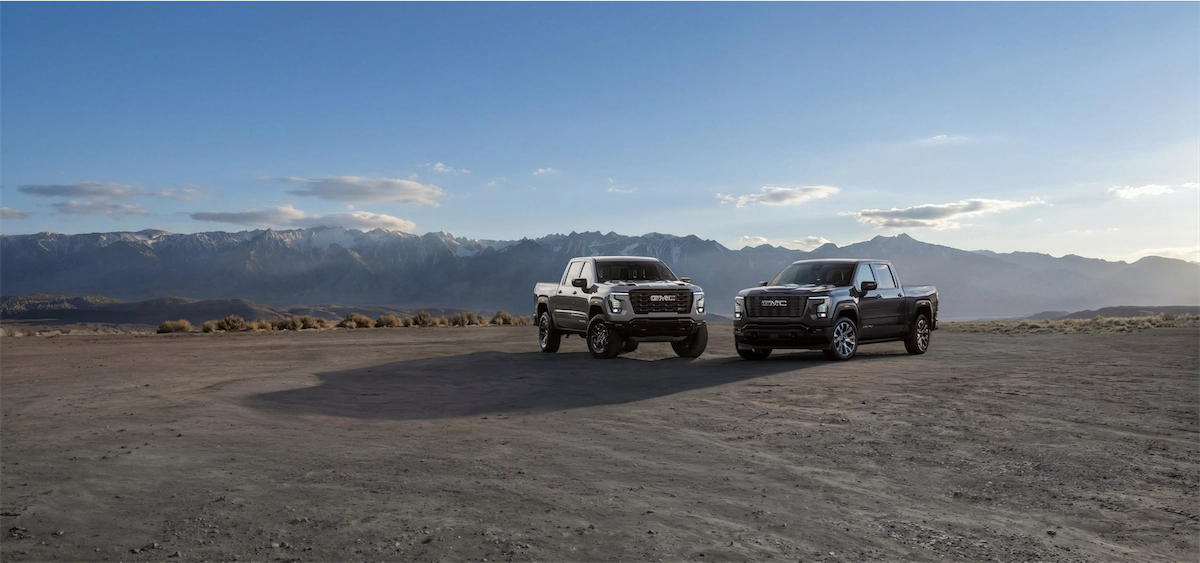 Two GMC pickups parked side by side on a desert flat with mountains behind