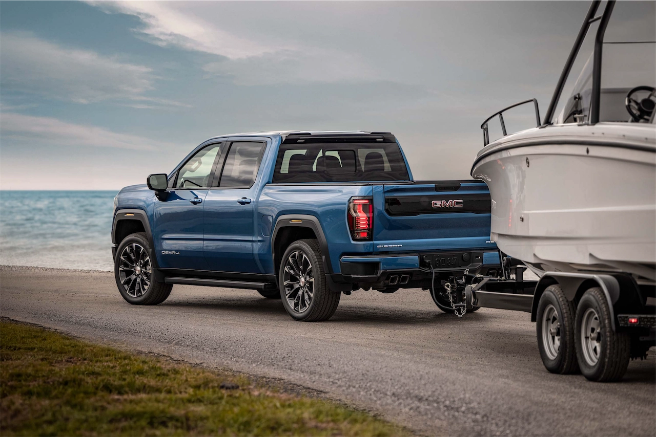 A blue GMC truck parked on a gravel path near the ocean, towing a white boat on a trailer.
