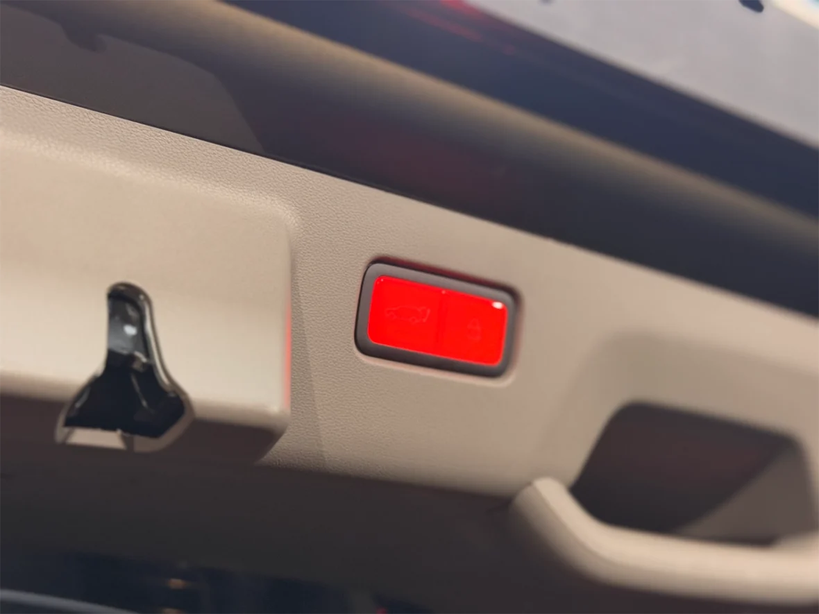 Close-up of a car trunk release button with a red light, located near a handle on a beige interior panel.