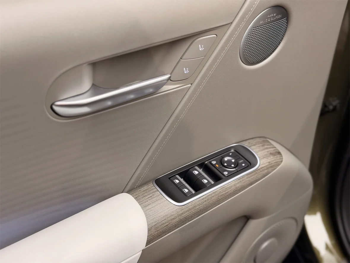 Interior view of a car door panel featuring a silver door handle, power window and seat controls, and a speaker, all against a beige background with wood accent.