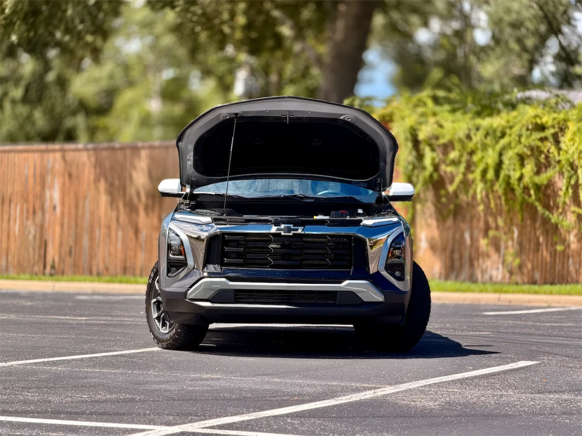 Front view of a Chevrolet SUV with the hood open, parked in a lot surrounded by greenery.