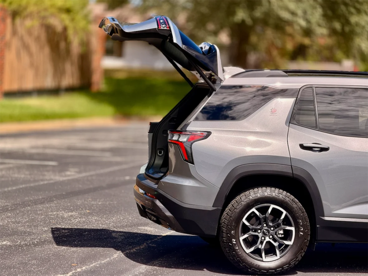 A silver SUV with its rear hatch open, parked on a paved surface with green grass and trees in the background.