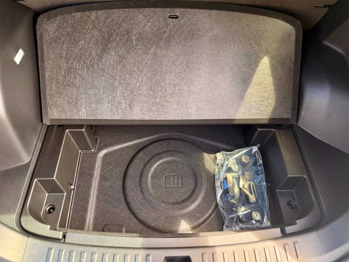 An empty car trunk compartment with a carpeted floor, featuring a circular indent and a plastic bag containing small items.