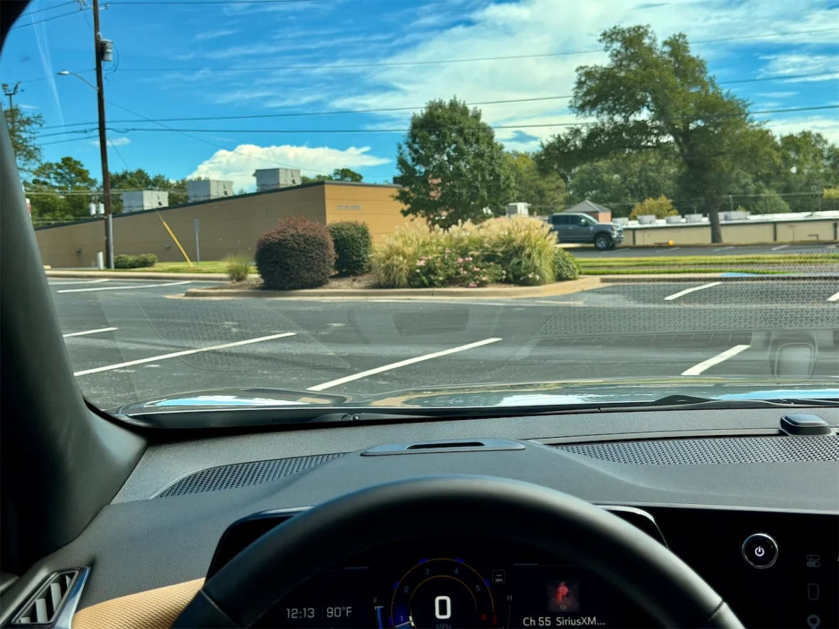View from inside a car showing the dashboard, steering wheel, and a parking lot scene with trees and a building in the background under a blue sky.