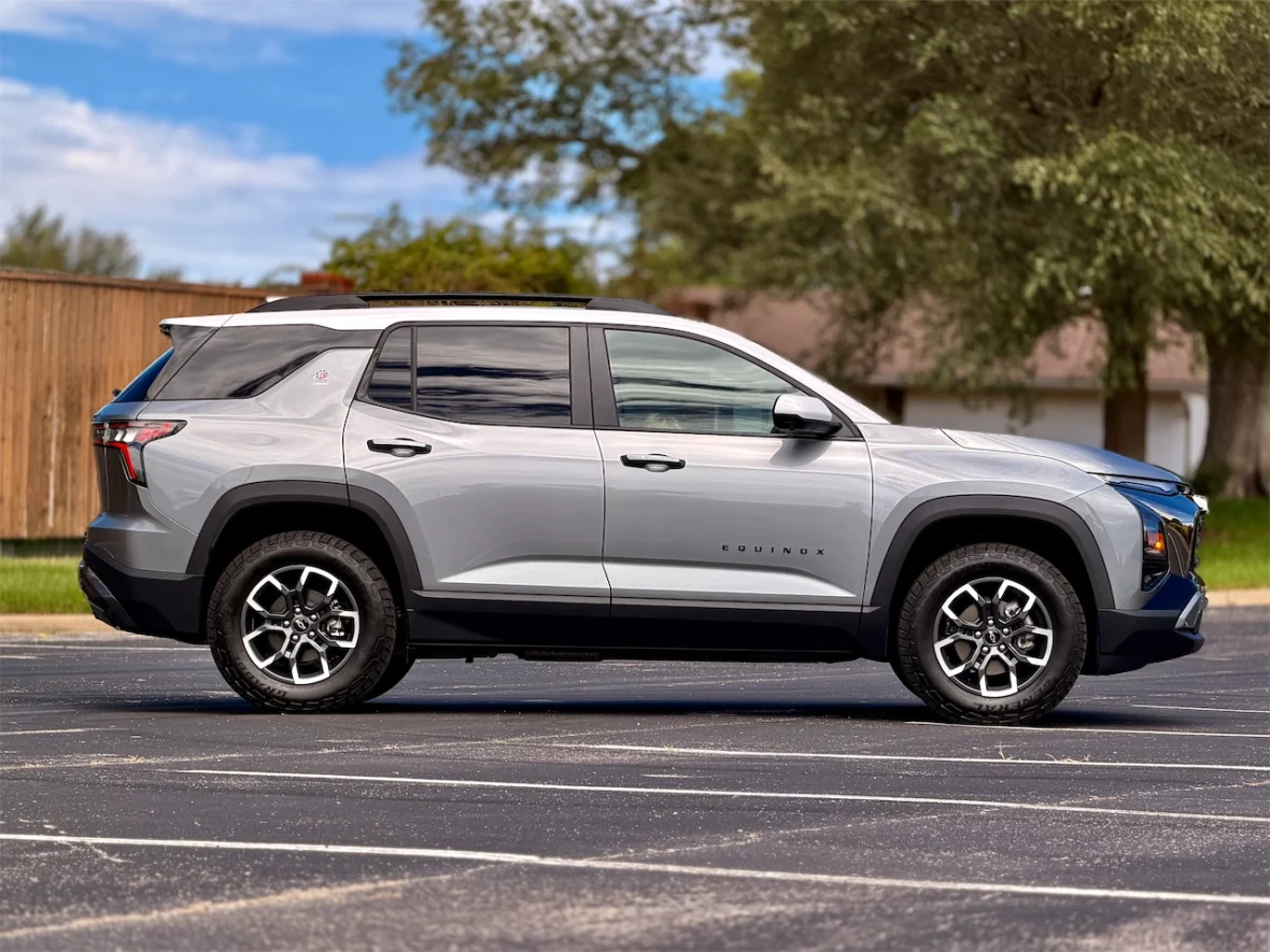 Side view of a gray Chevrolet Equinox parked on a paved surface with trees and a wooden fence in the background.