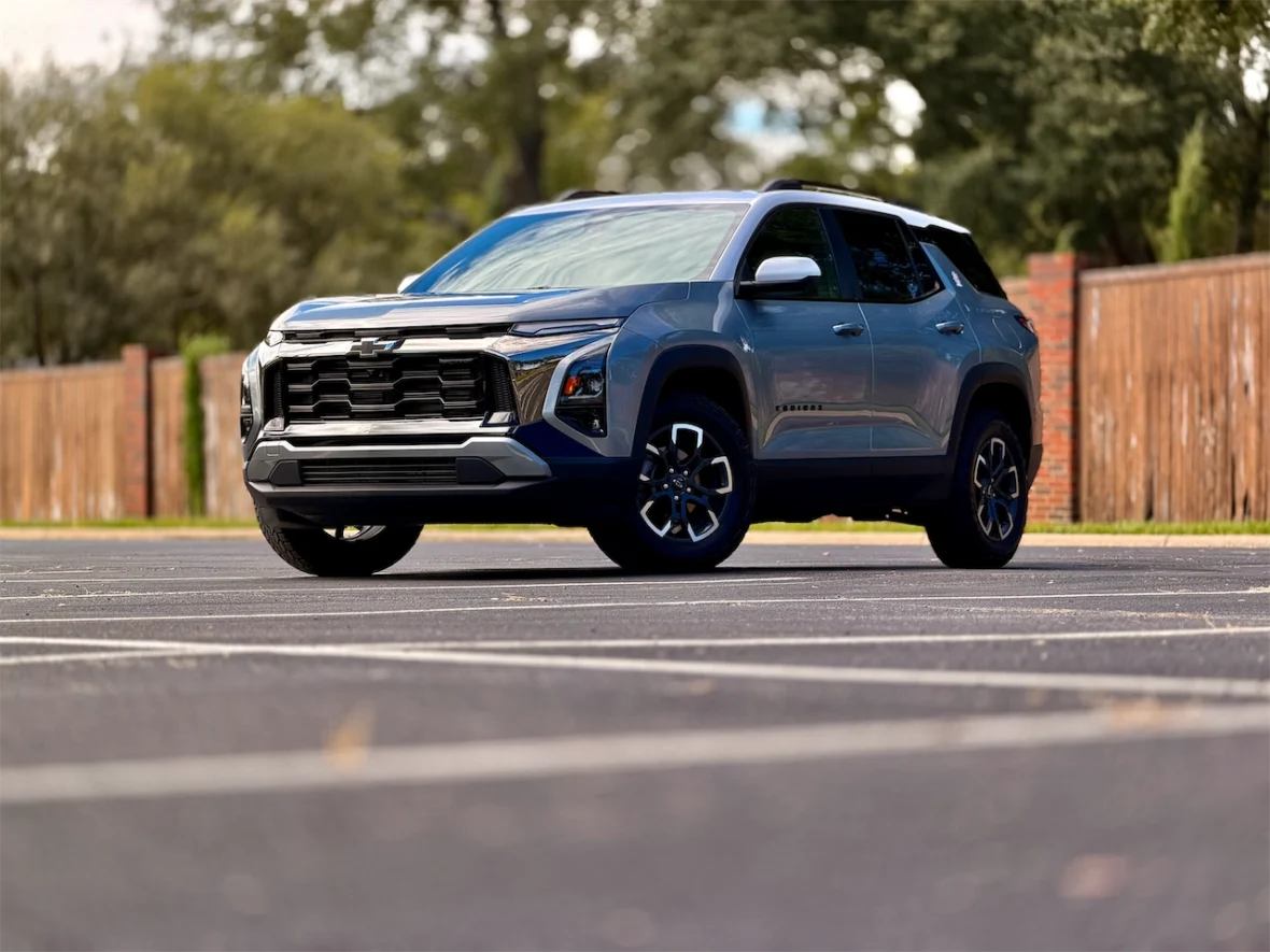 A silver and blue Chevrolet SUV parked in an empty lot, with trees and a wooden fence in the background.