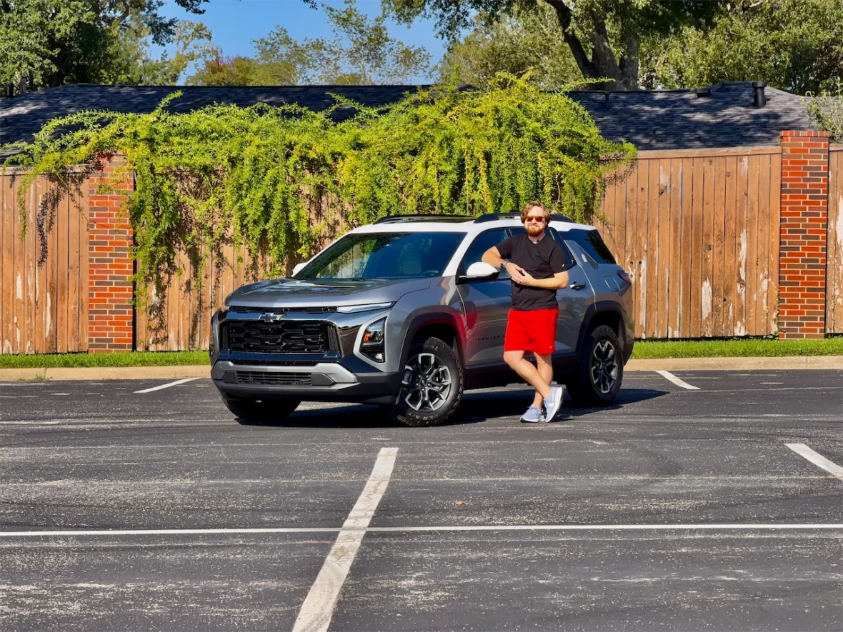 A man in a black shirt and red shorts stands casually next to a gray Chevrolet SUV parked in an empty lot, with a wooden fence and greenery in the background.