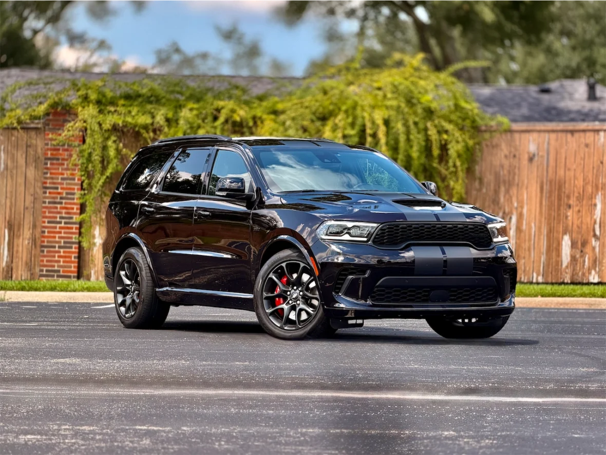 A black SUV parked on a paved surface with greenery and a wooden fence in the background.