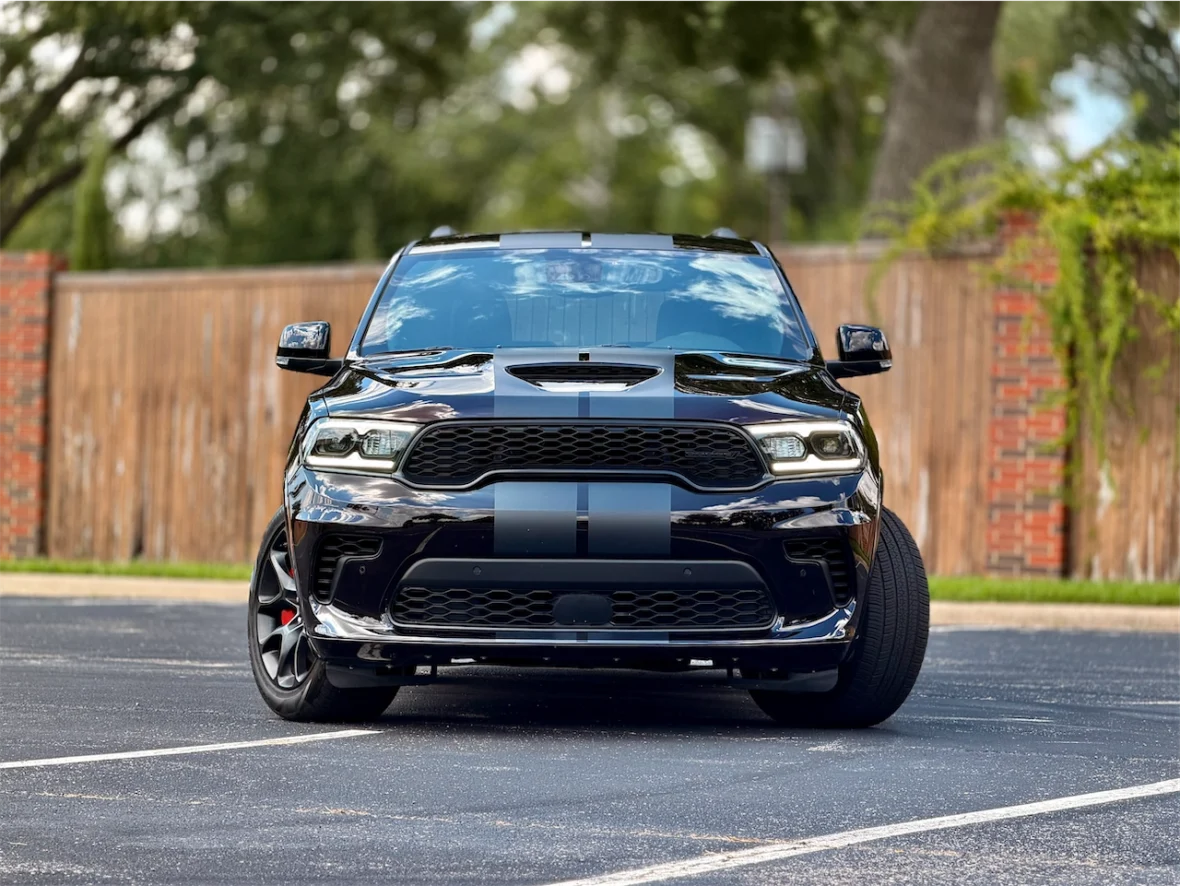 Front view of a black sports SUV with a hood scoop and racing stripes, parked in a lot with a wooden fence in the background.