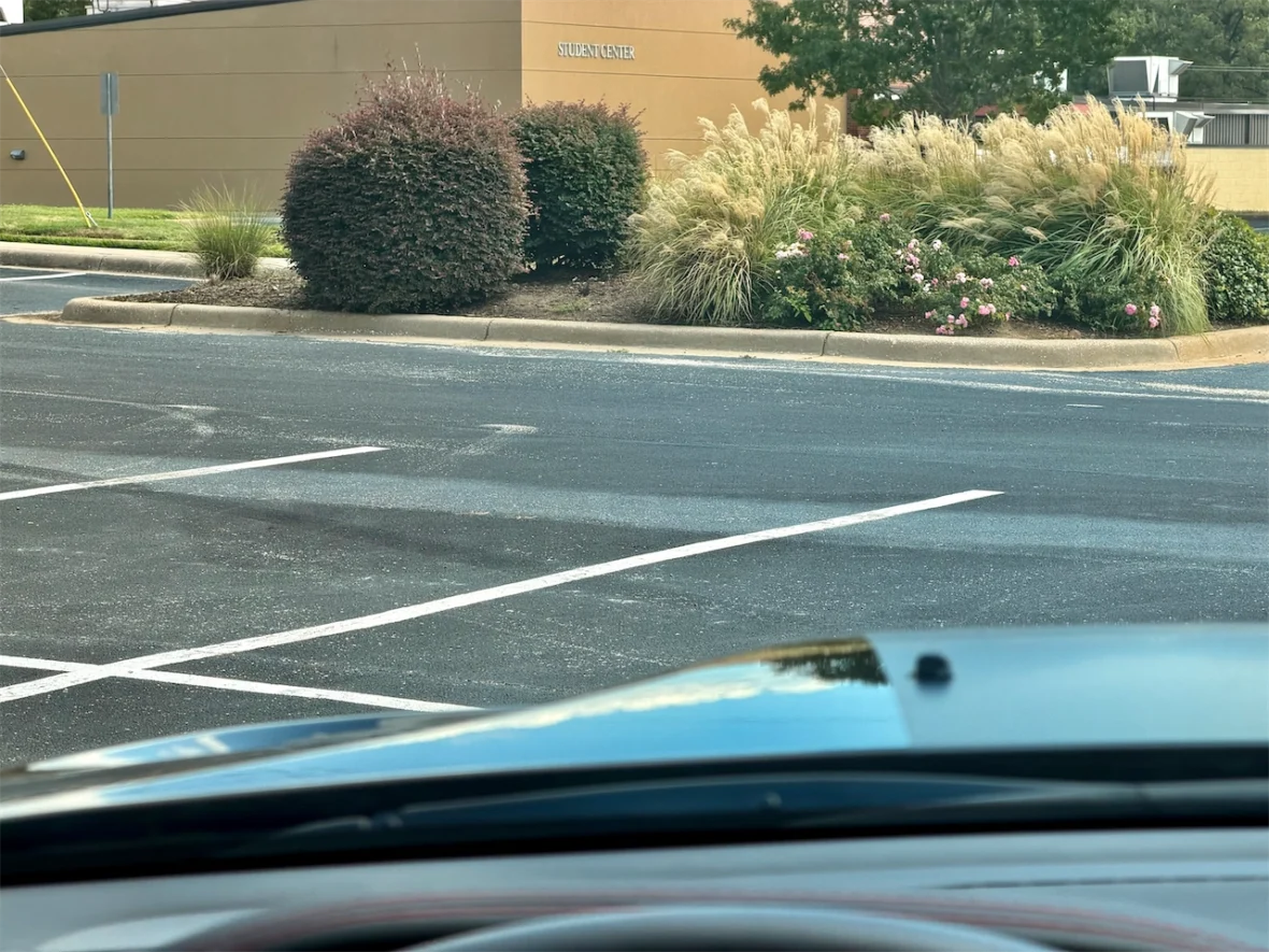 View of a partially empty parking lot with marked spaces, shrubs, and decorative plants in the foreground, and a building in the background labeled 'Student Center'.