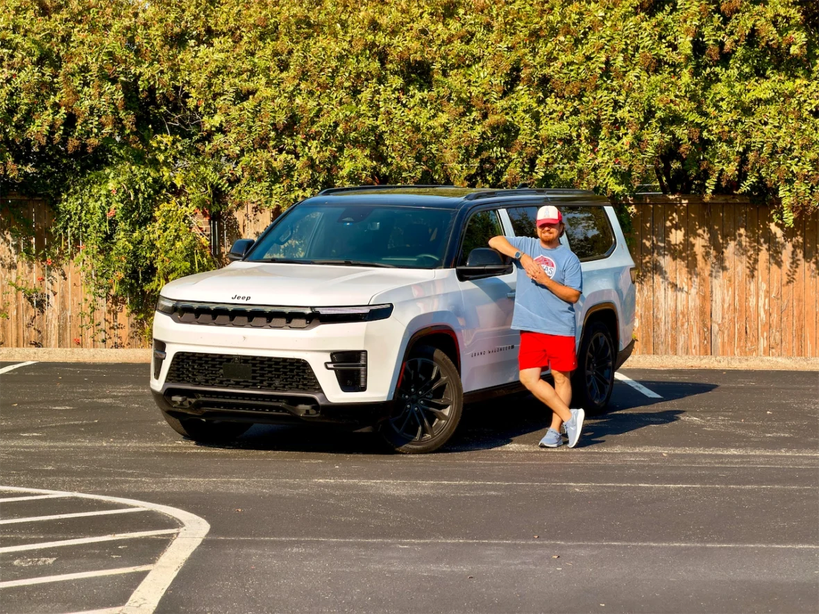 A man in a blue t-shirt and red shorts stands next to the 2026 Jeep Grand Wagoneer L parked in a lot with greenery in the background.