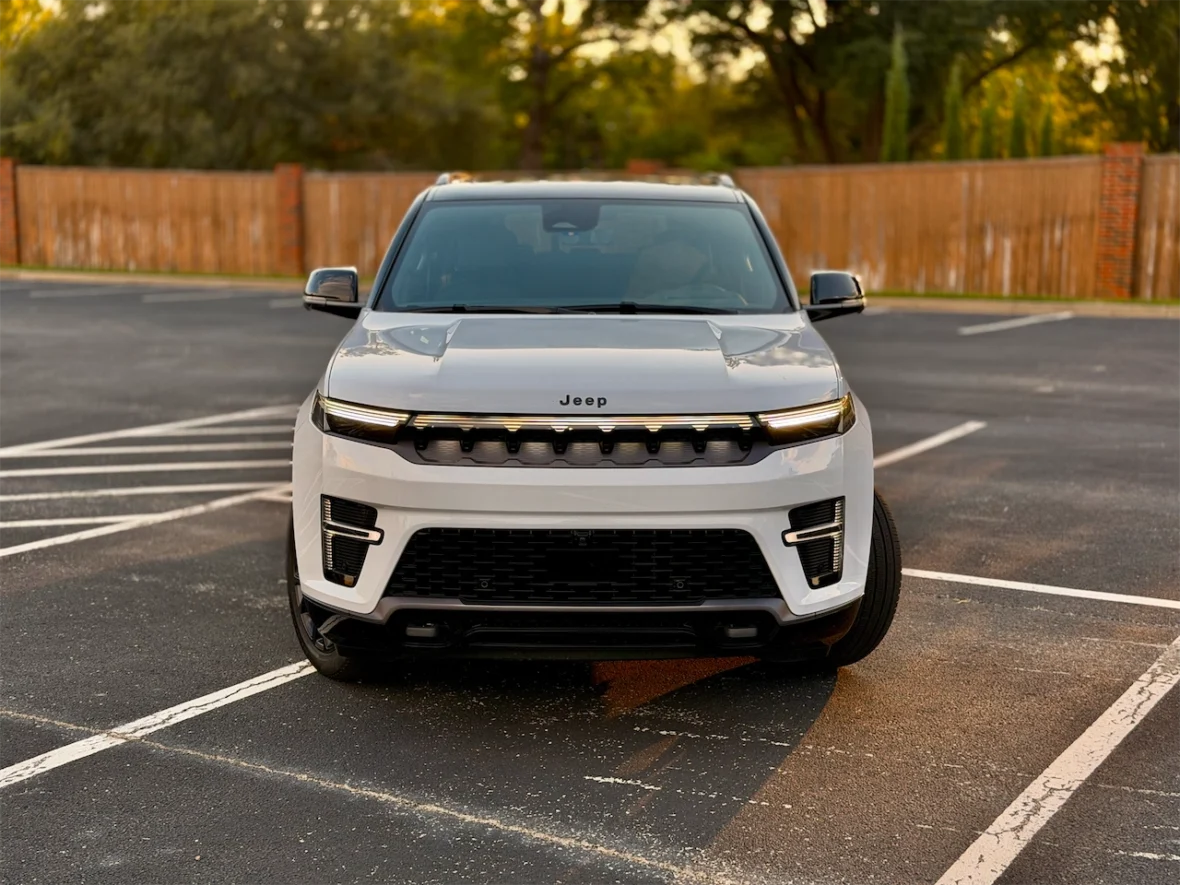 Front view of the 2026 Jeep Grand Wagoneer L parked in a lot with a blurred background of trees and a wooden fence.