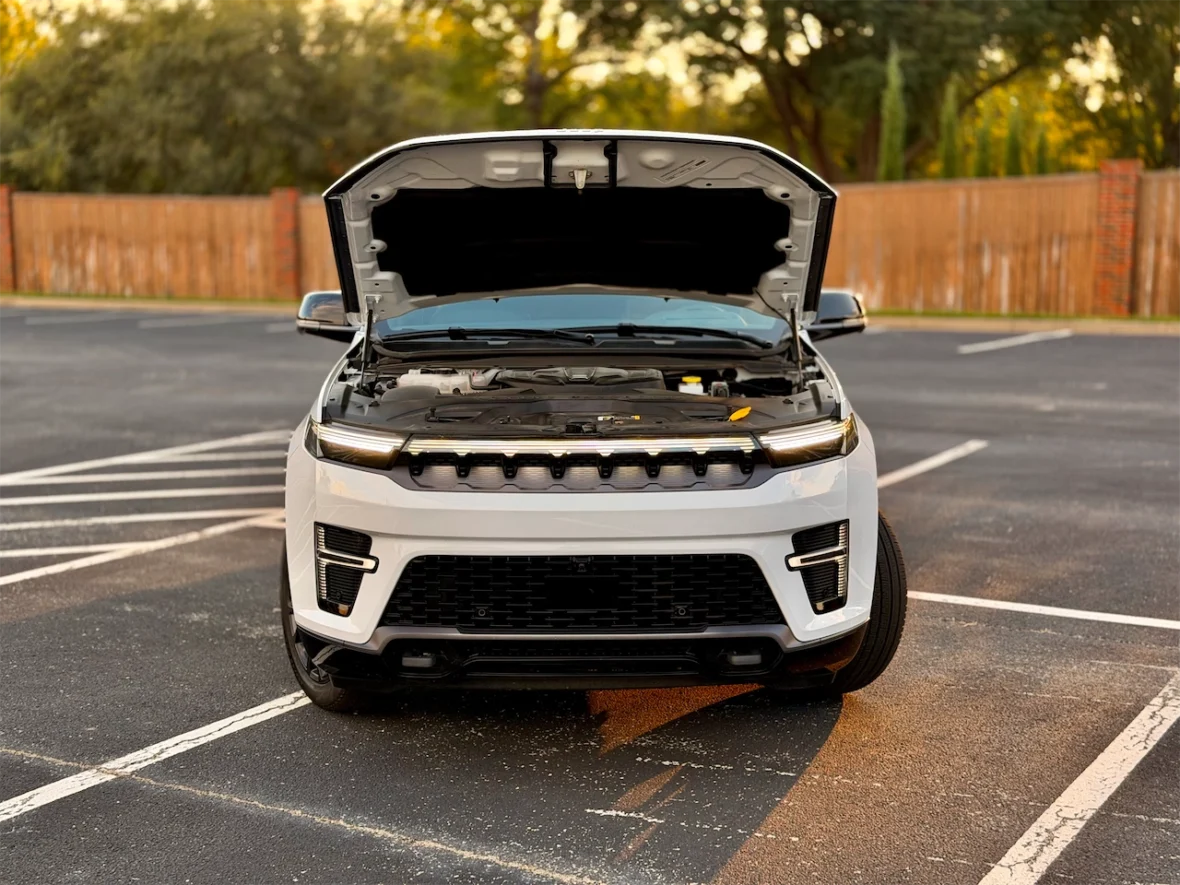 Front view of the 2026 Jeep Grand Wagoneer L with the hood open, showcasing the engine compartment, parked in a lot with a blurred wooden fence in the background.