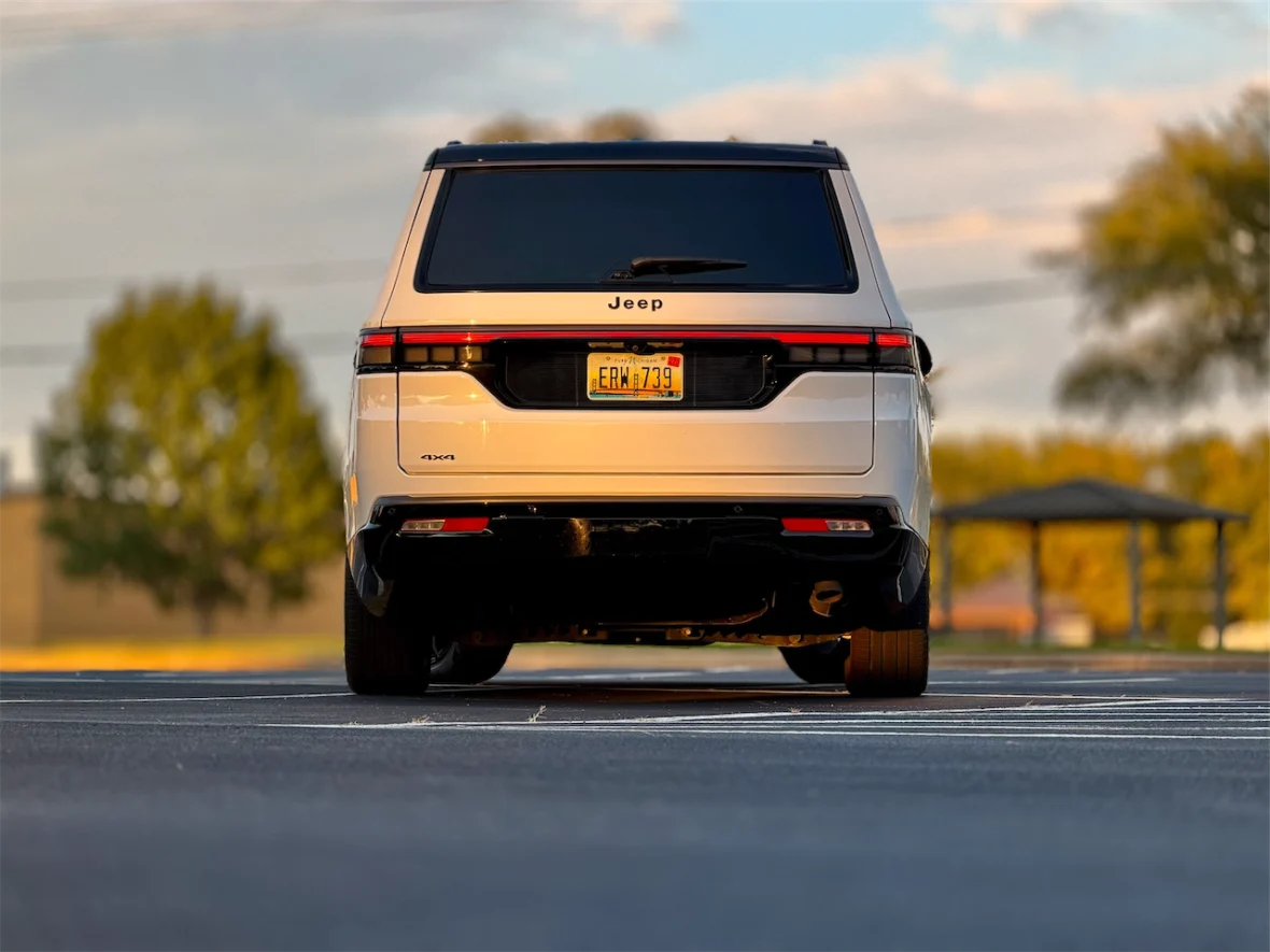 Rear view of the 2026 Jeep Grand Wagoneer L parked on a blacktop surface with trees and a cloudy sky in the background.