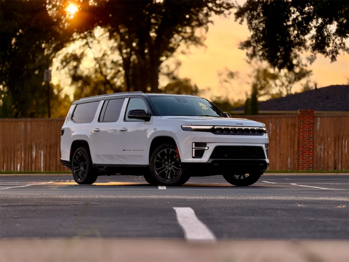 The 2026 Jeep Grand Wagoneer L parked on a road during sunset, with a blurred background of trees and a wooden fence.