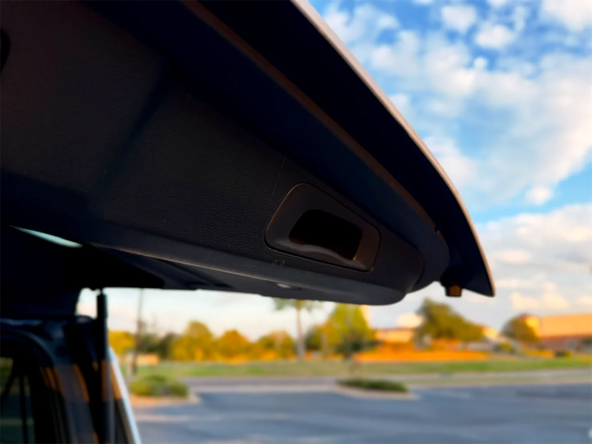 Close-up of a car's sunroof opening mechanism, with a blurred background of trees and a parking lot.