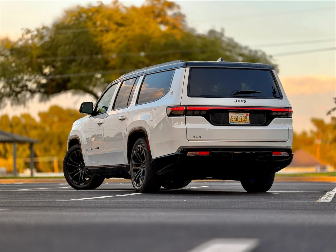 The 2026 Jeep Grand Wagoneer L parked in an empty lot, viewed from the rear, with trees and a sunset in the background.
