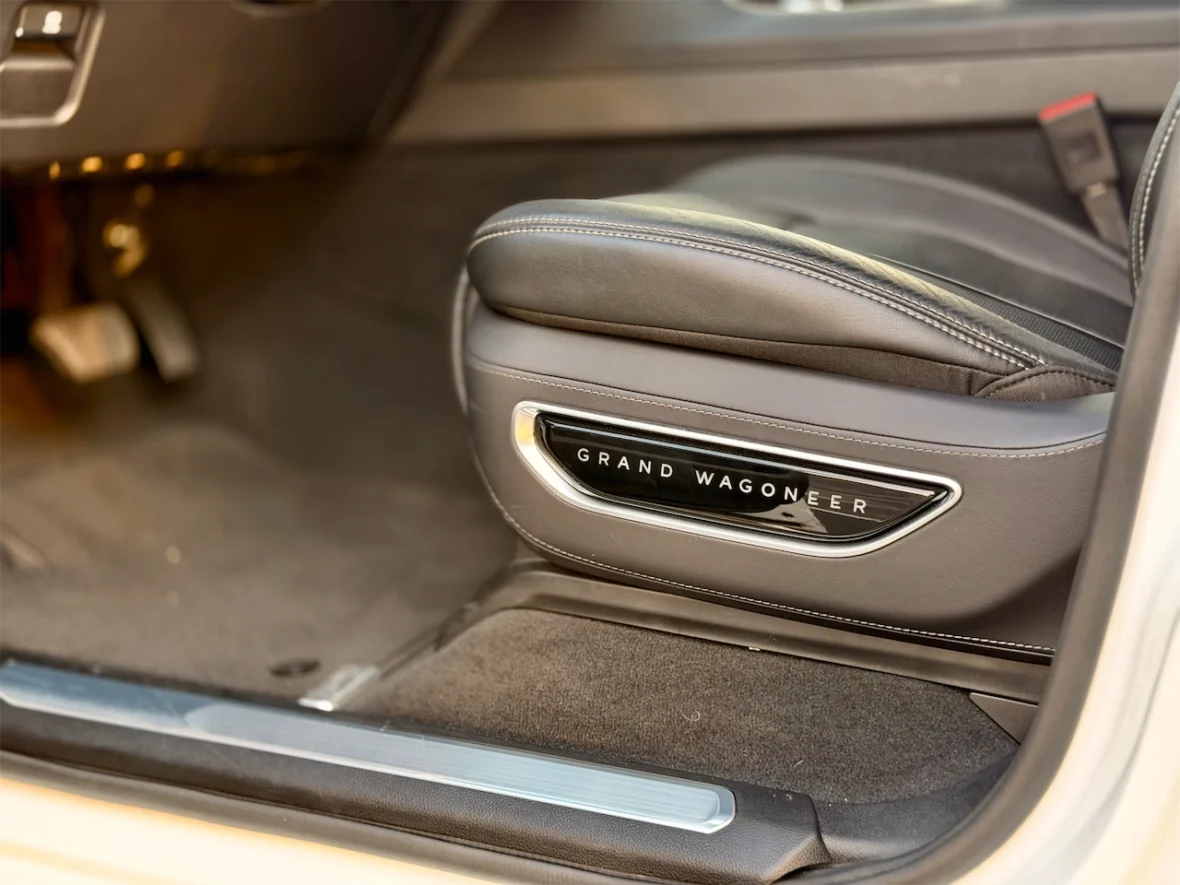 Interior view of a Grand Wagoneer showing the driver's seat, featuring black leather upholstery and an elegant trim with a logo.