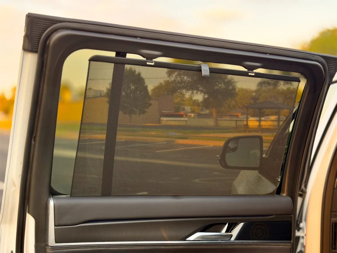 A close-up view of a vehicle's side window equipped with a retractable mesh sunshade, partially extended, showing a blurred outdoor background.