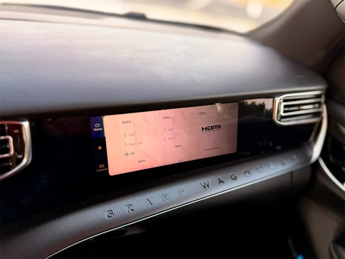 Close-up of a car dashboard featuring a touchscreen display with audio and video selection options, including USB and Bluetooth connectivity. The name 'GRAND WAGONEER' is visible on the dashboard surface.