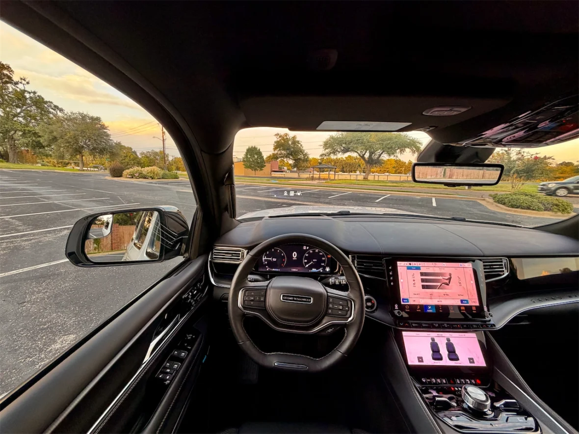 Interior view of a luxury car showcasing the dashboard, steering wheel, and large touchscreen display, with a clear view outside showing a parking lot and trees.