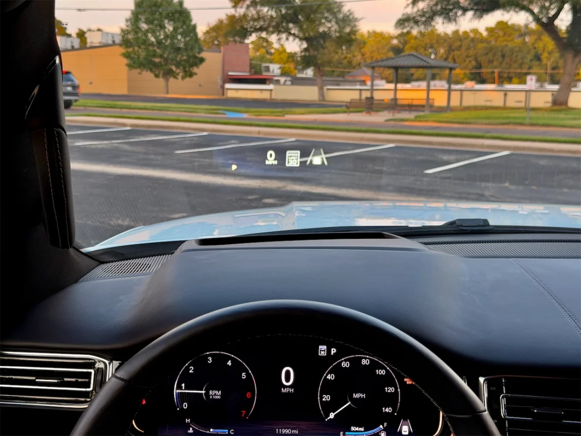 Interior view of a vehicle's dashboard showing the speedometer, a head-up display with speed and navigation indicators, and an exterior view of a parking area.