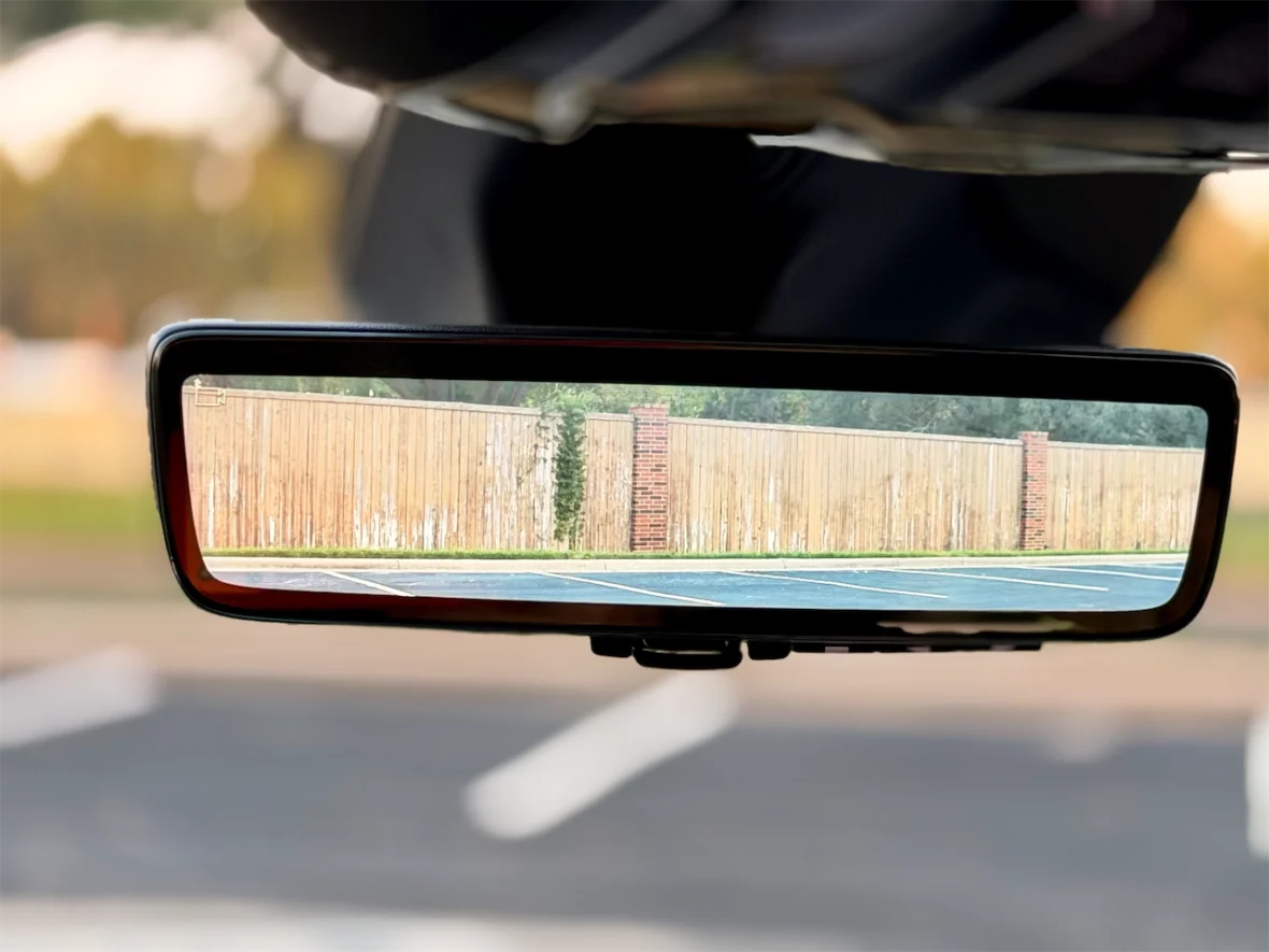 View of a wooden fence captured in a rearview mirror inside a car, with an asphalt parking lot visible in the background.
