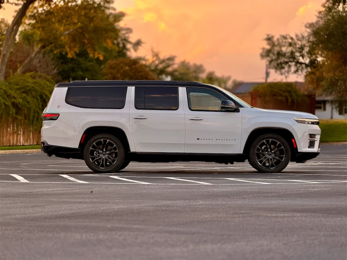 A white Grand Wagoneer SUV parked in an empty lot, with a sunset background and trees in the distance.