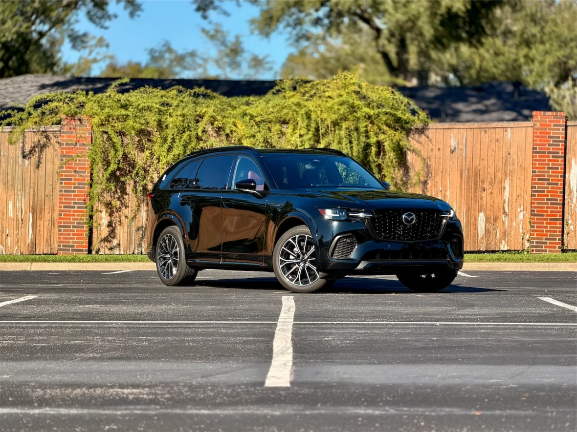 A black Mazda SUV parked in an empty parking lot with a wooden fence and greenery in the background.