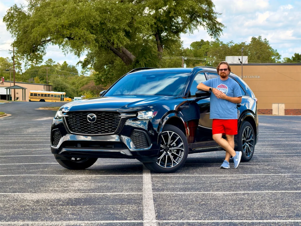 A man leaning against a black Mazda SUV in a parking lot, wearing a gray t-shirt and red shorts.