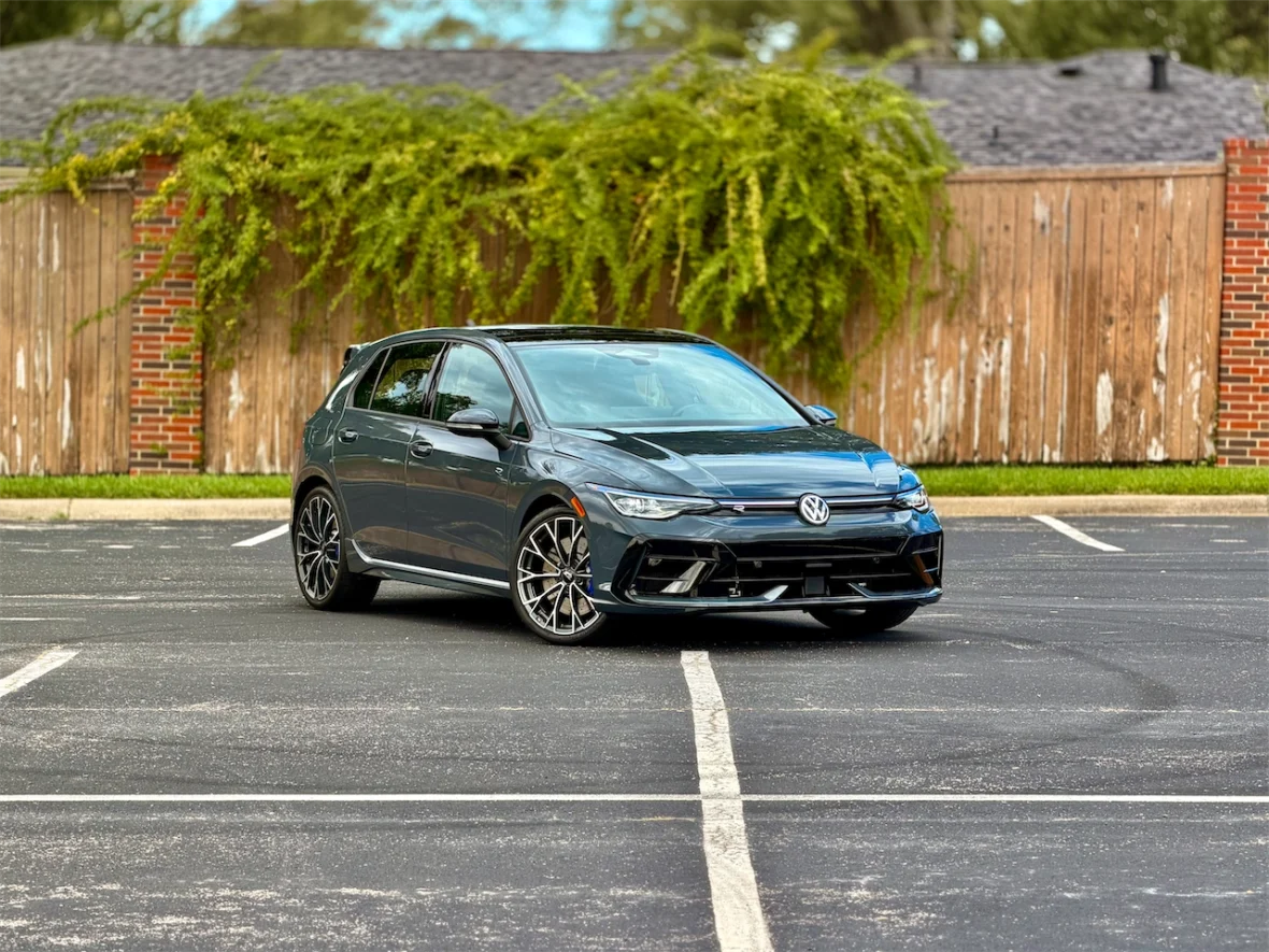 A Volkswagen hatchback parked diagonally in an empty parking lot with greenery and a wooden fence in the background.