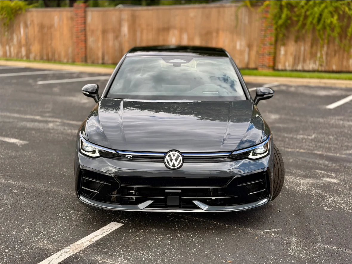 Front view of a black Volkswagen car parked on a paved surface with a wooden fence in the background.