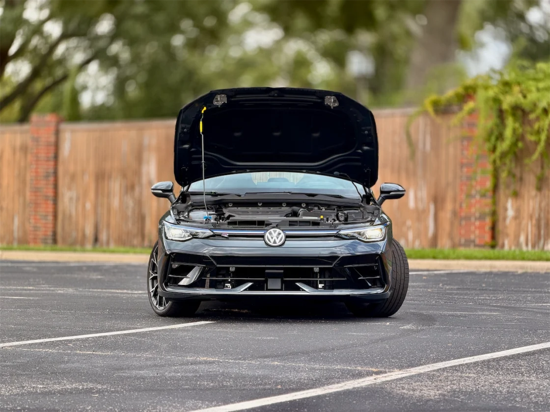 Front view of a black Volkswagen car with the hood open, showcasing the engine, parked in a lot with a wooden fence in the background.