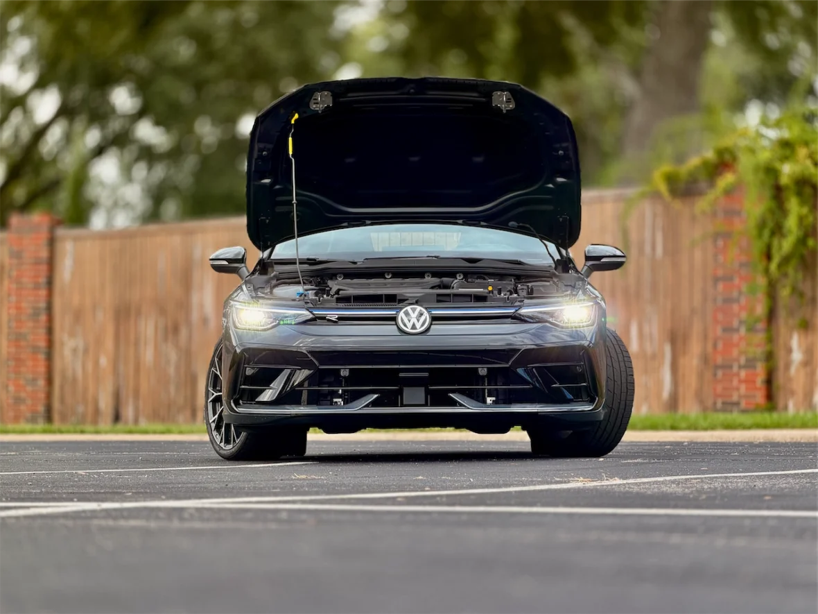 Front view of a black Volkswagen car with the hood open, showcasing the engine compartment, parked on a paved surface with a wooden fence in the background.