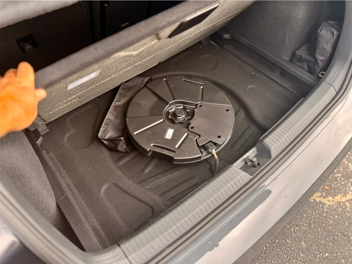 View of a car trunk with a round black storage compartment and a hand reaching towards it.