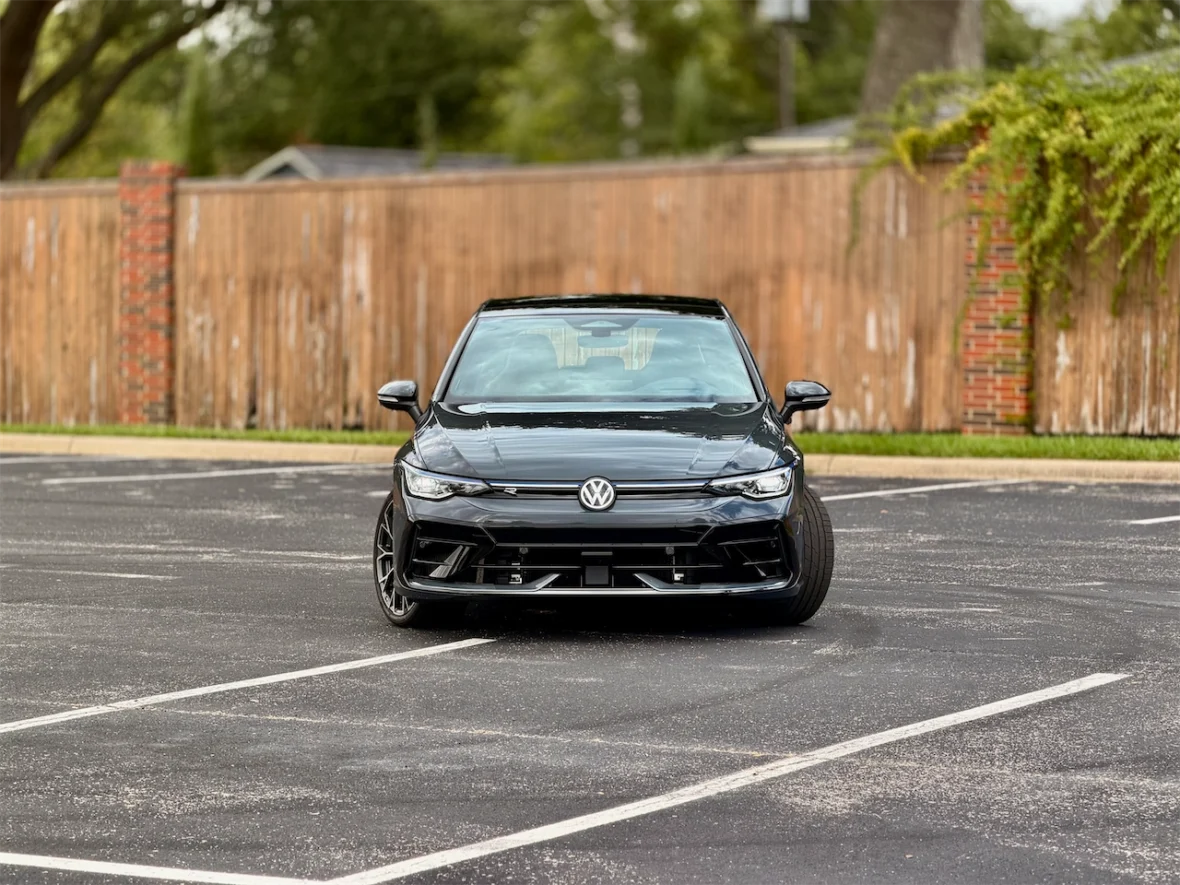 A black Volkswagen car parked in a lot, facing the camera, with a wooden fence in the background.