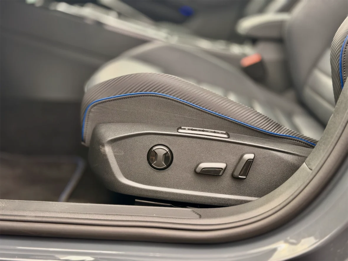 Close-up of a car seat adjuster with buttons for seat height and backrest angle, featuring a gray fabric with blue stitching.