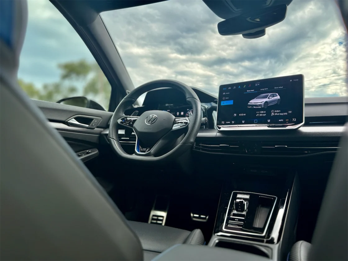 Interior view of a modern Volkswagen car dashboard, featuring a steering wheel, digital display screen, and gear shift, against a cloudy sky in the background.