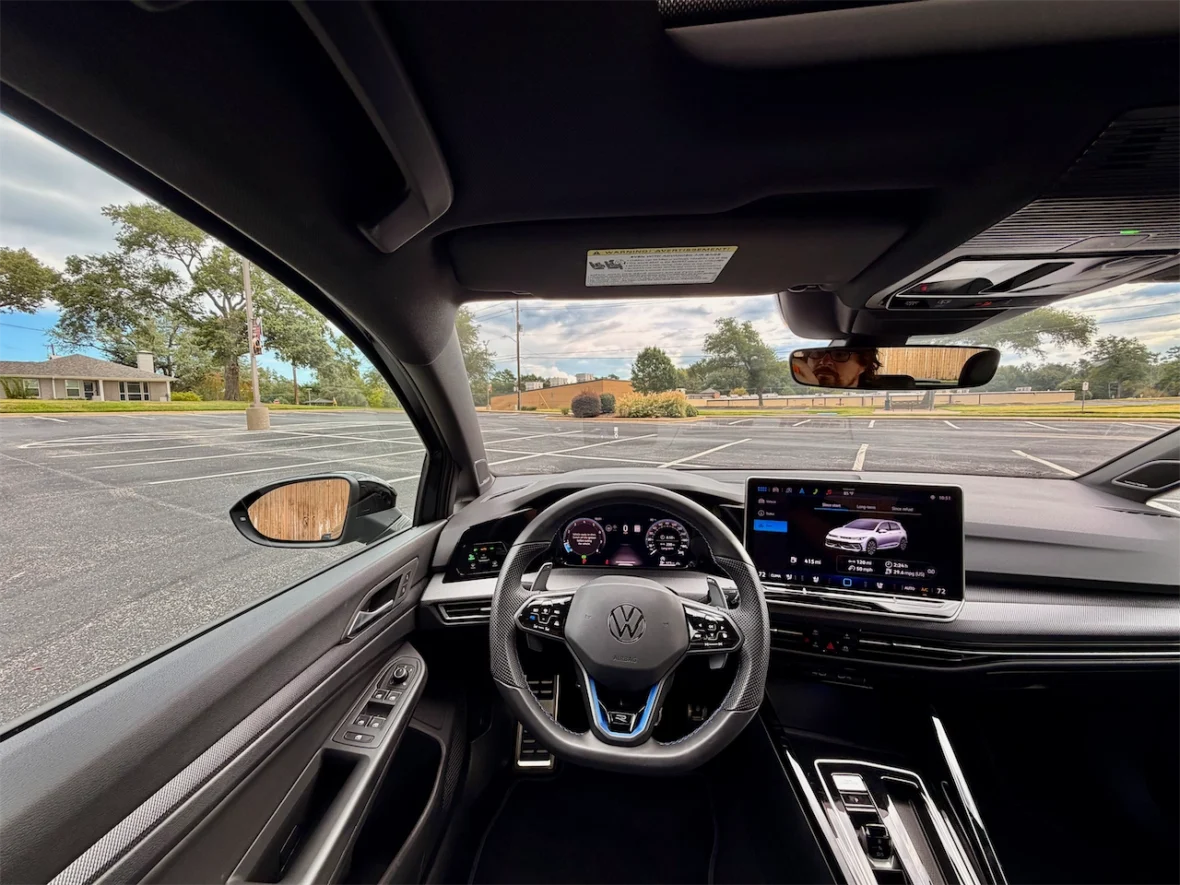 Interior view of a Volkswagen car, showing the steering wheel, dashboard, and infotainment screen, with an empty parking lot visible through the windshield.