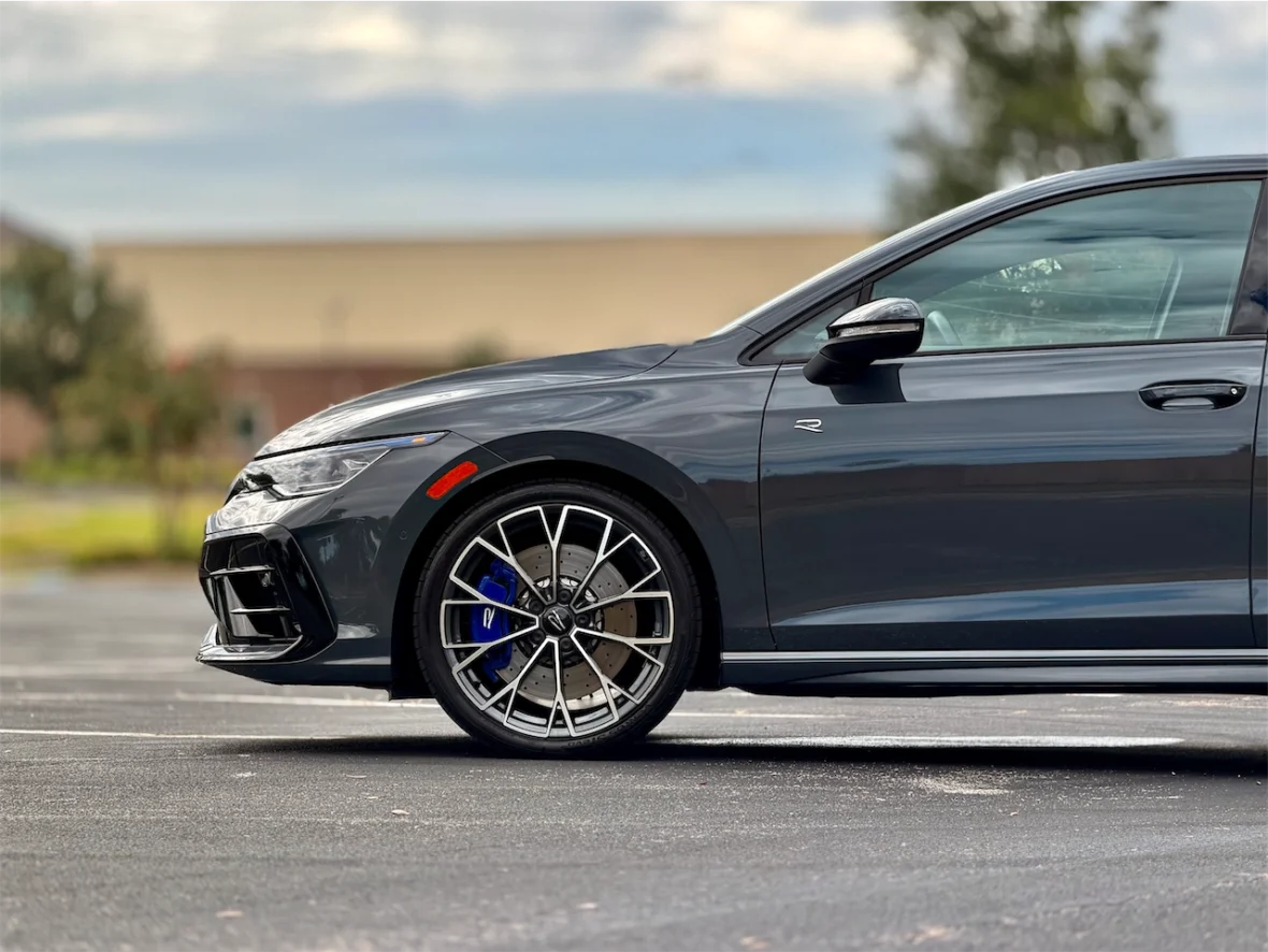 Side view of a sleek gray car highlighting a stylish alloy wheel with blue brake calipers, parked on an asphalt surface with blurred greenery in the background.