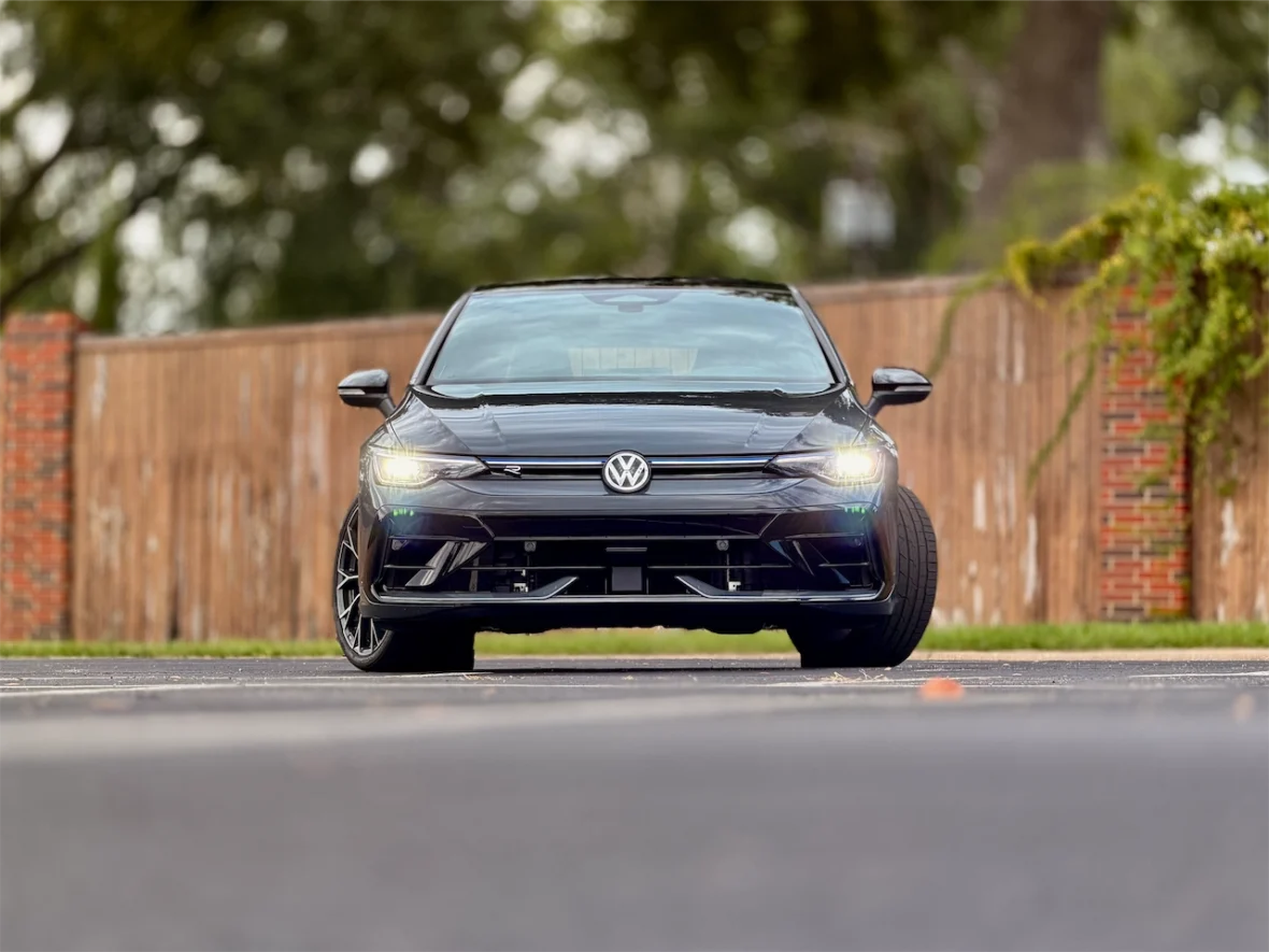 Front view of a black Volkswagen car on a road with a wooden fence in the background.