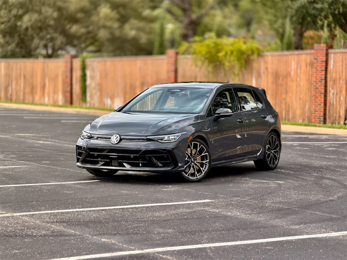 A gray Volkswagen hatchback parked in an empty lot with a wooden fence in the background.