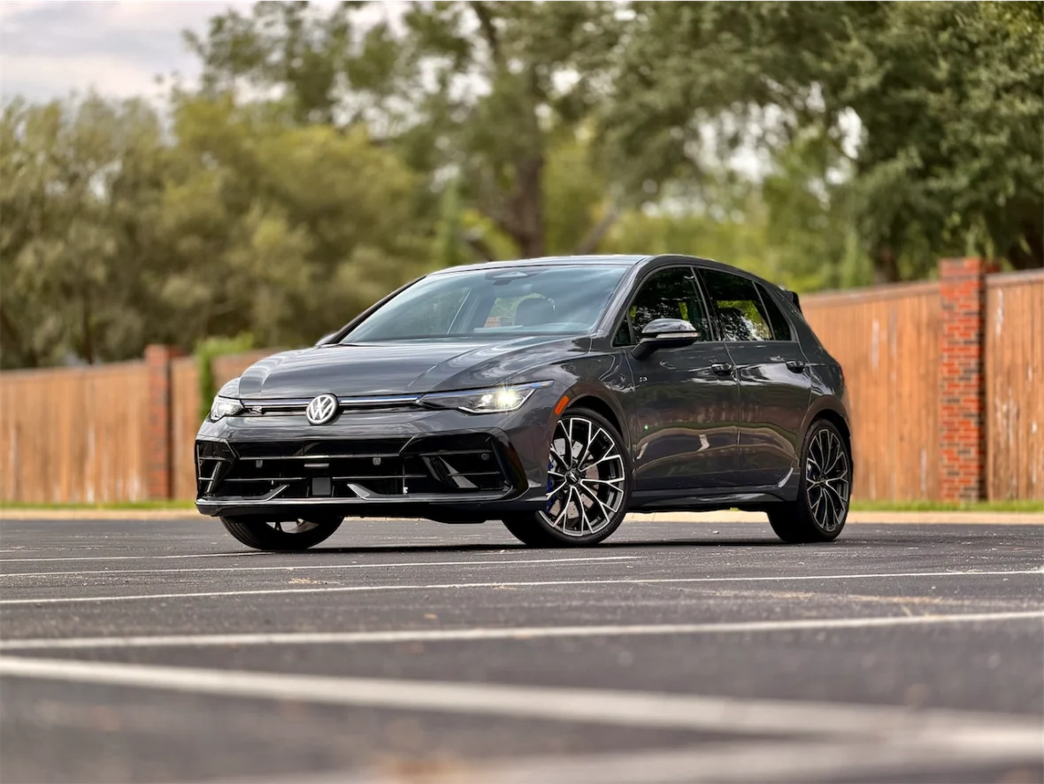 A sleek black Volkswagen hatchback parked in an empty lot with a wooden fence and trees in the background.