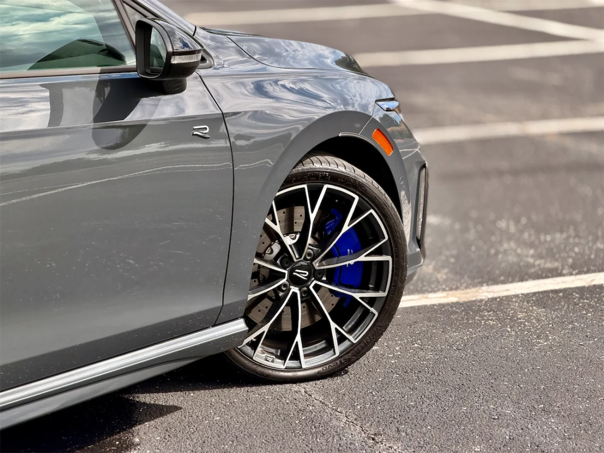 Close-up of a gray car's front wheel and side view, showcasing a stylish alloy rim with blue accents and a visible brake caliper.