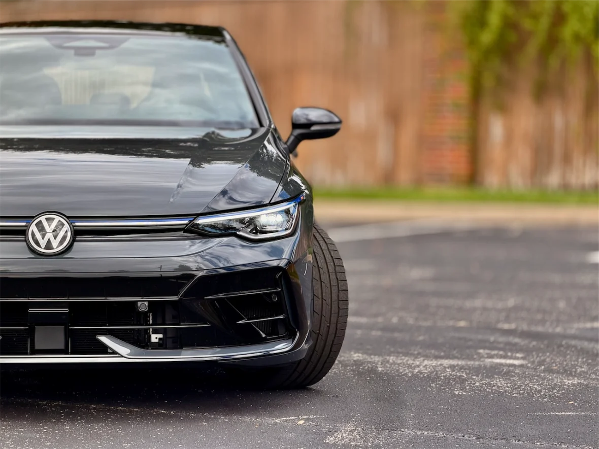 Front view of a black Volkswagen car parked on asphalt with a blurred wooden fence in the background.