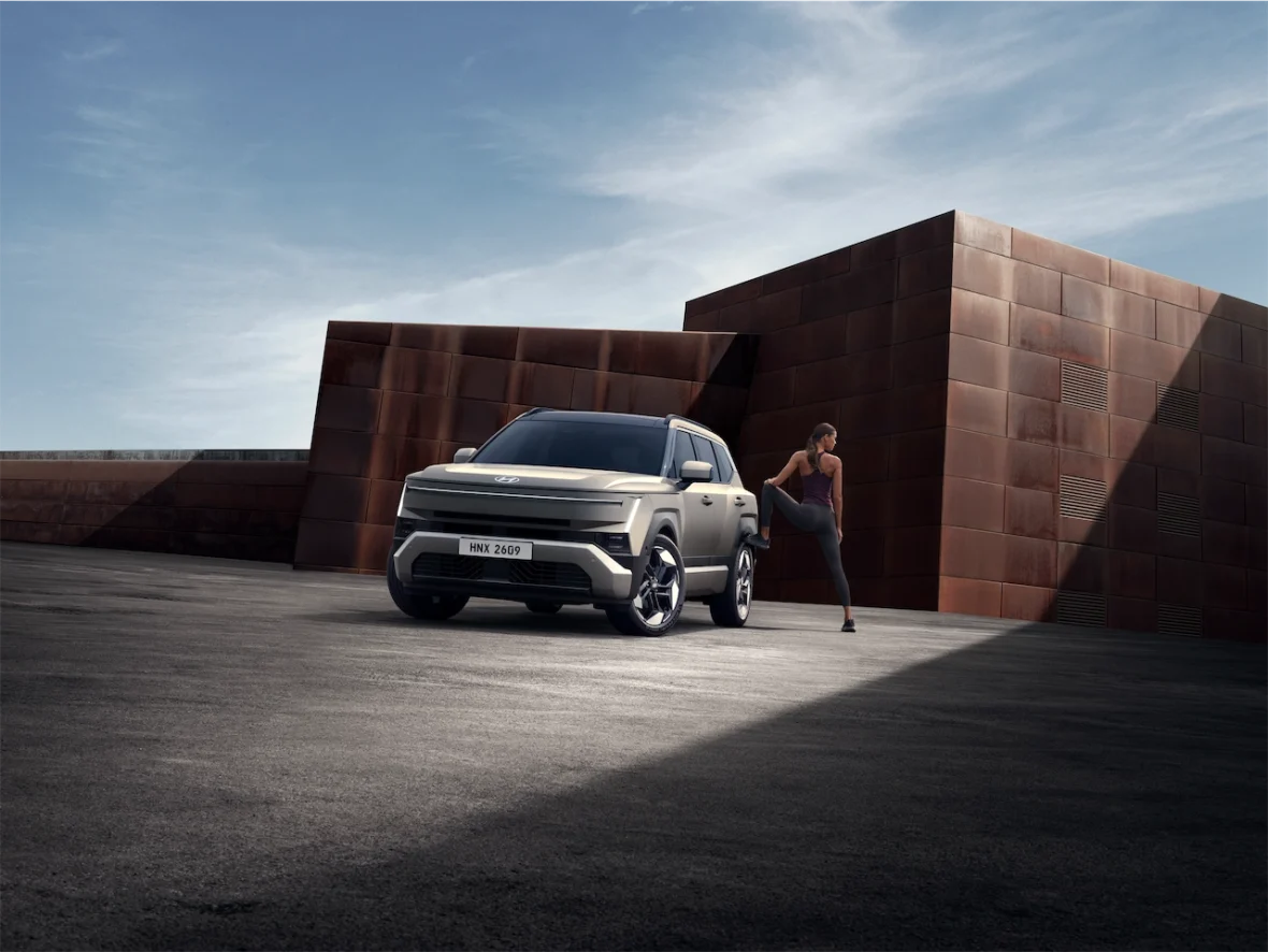 A modern SUV parked on a textured surface, with a person in athletic wear stretching next to it against a backdrop of angular, rust-colored architecture and a clear sky.