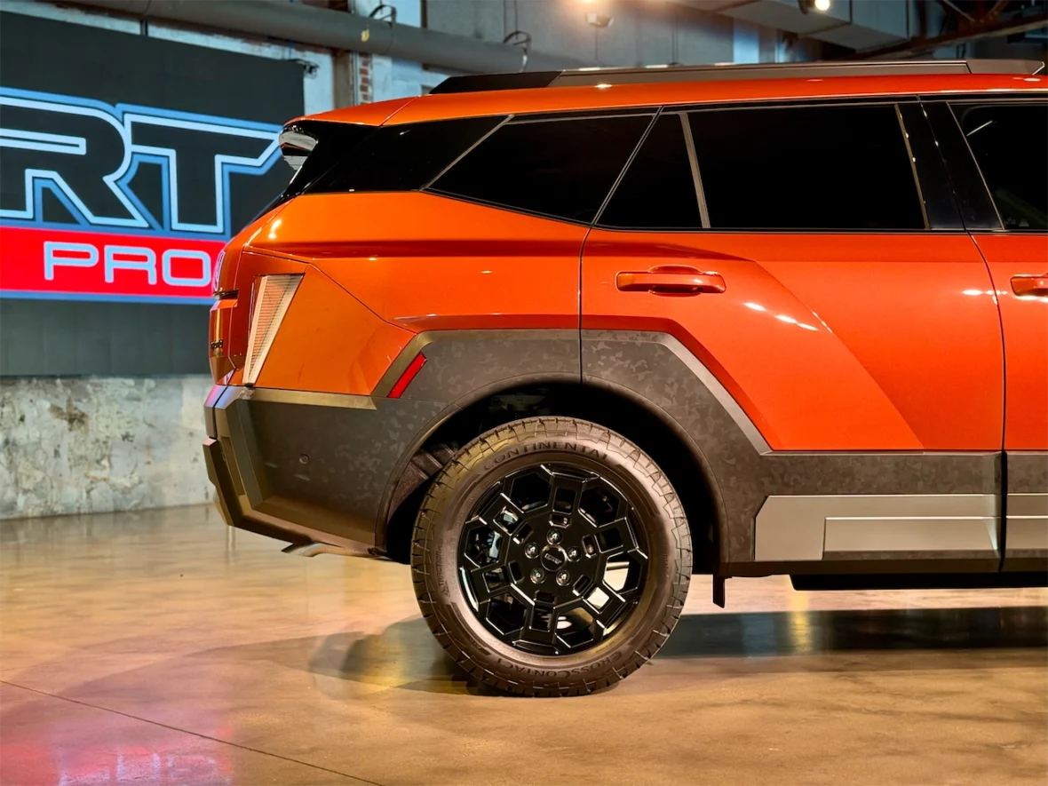 Side view of a modern orange SUV with a bold design, showcasing its black rims and detailed exterior features, set against a backdrop with illuminated branding.