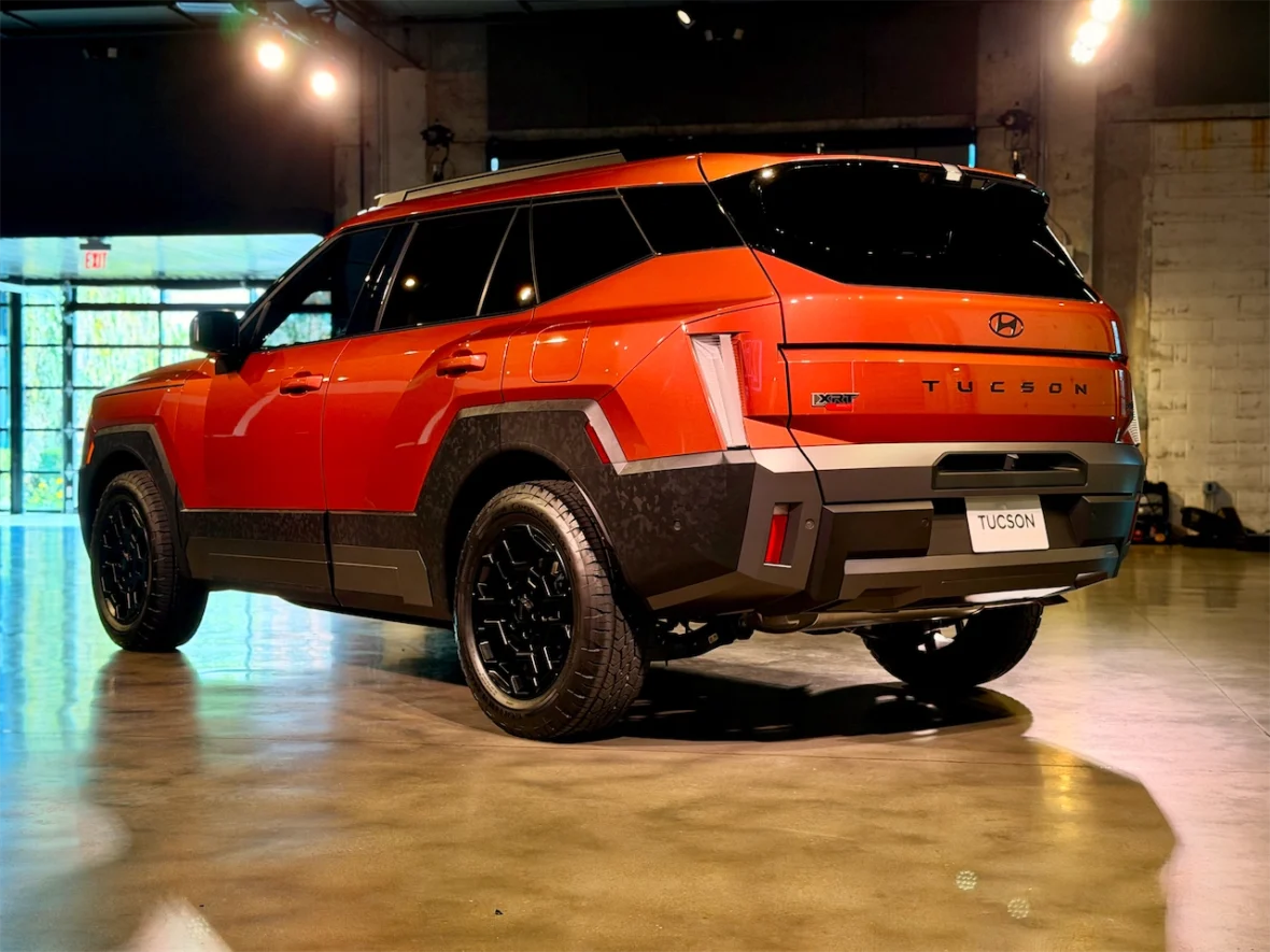 A rear view of a vibrant orange Hyundai Tucson SUV displayed indoors, showcasing its sleek design and black alloy wheels under soft lighting.