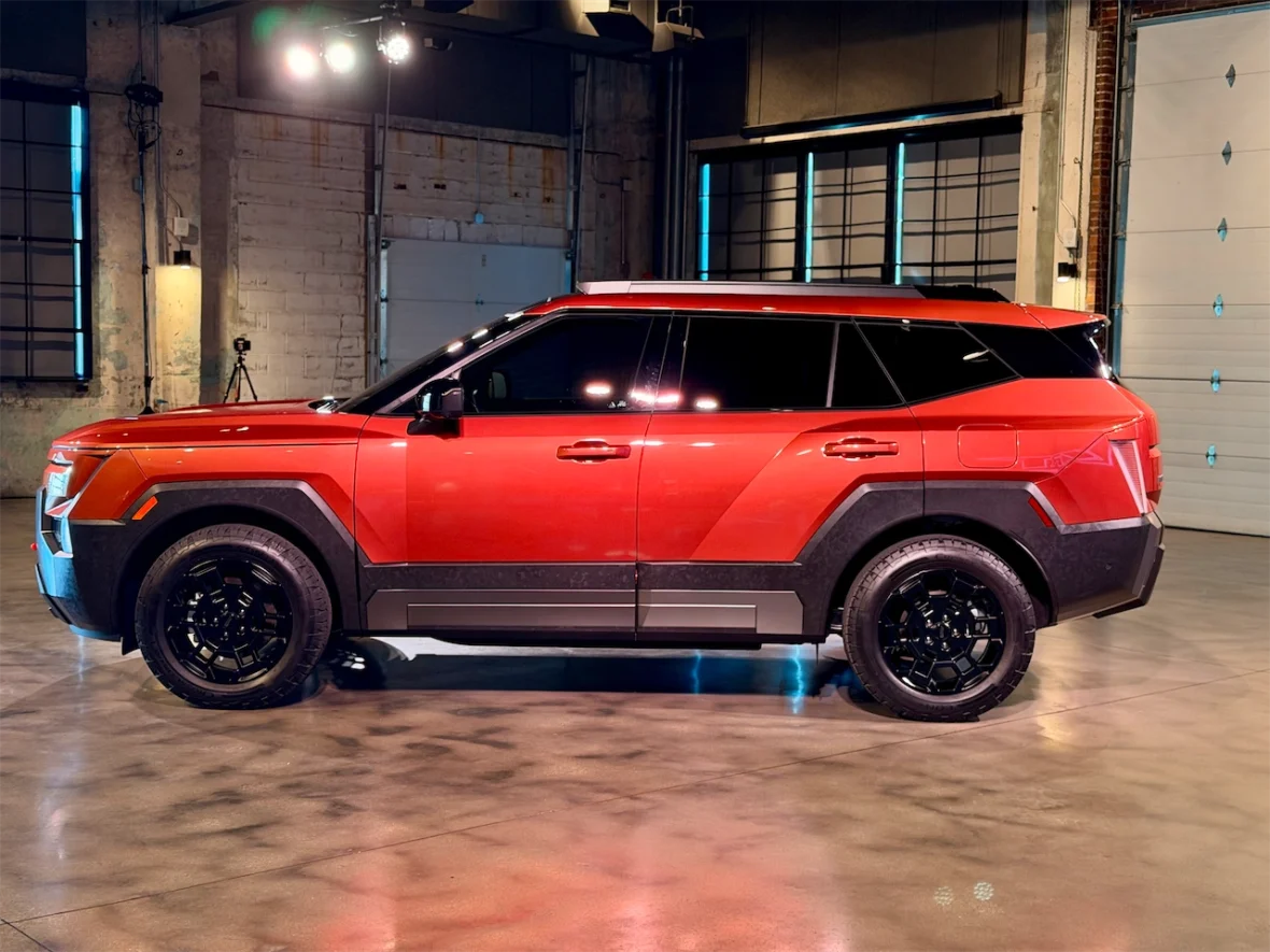 A side view of a red SUV with a modern design, featuring black accents and wheels, showcased in an industrial setting with spotlighting.