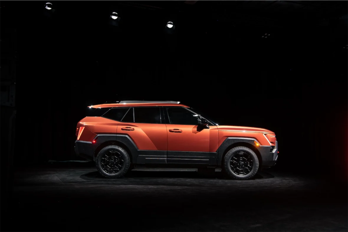 A side view of a modern orange SUV parked under studio lights against a dark background.